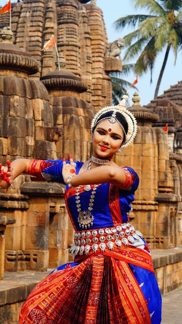 Eine Frau in einem blauen und roten Sari tanzt vor einem mit Flaggen geschmückten Tempel, umgeben von Bäumen unter einem klaren blauen Himmel.