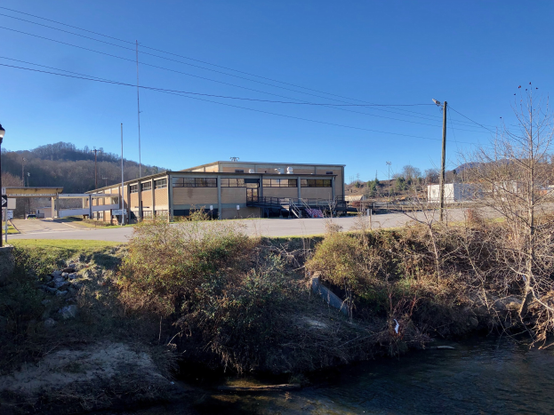 Ein Fluss fließt durch ein grünes Feld neben einem Gebäude in der Nähe von Bäumen, Pflanzen, Gras, Steinen, Straßenlaternen, Strommasten, Stromkabeln, Hügeln und einem klaren blauen Himmel, wobei das Gebäude als Standort einer geplanten Wasseraufbereitungsanlage geplant ist.