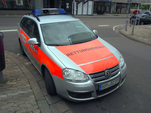 Ein Polizeiwagen steht am Straßenrand neben einem Mülleimer auf dem Gehweg, mit Gebäuden, Polen, Brettern, Pflanzen, Bäumen und anderen Objekten im Hintergrund.