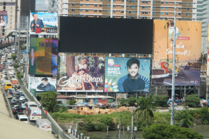 Eine belebte Stadtstraße mit starkem Verkehr, hohen Gebäuden und einem prominenten Werbeplakat, umgeben von Grün, Wasser und Strommasten.