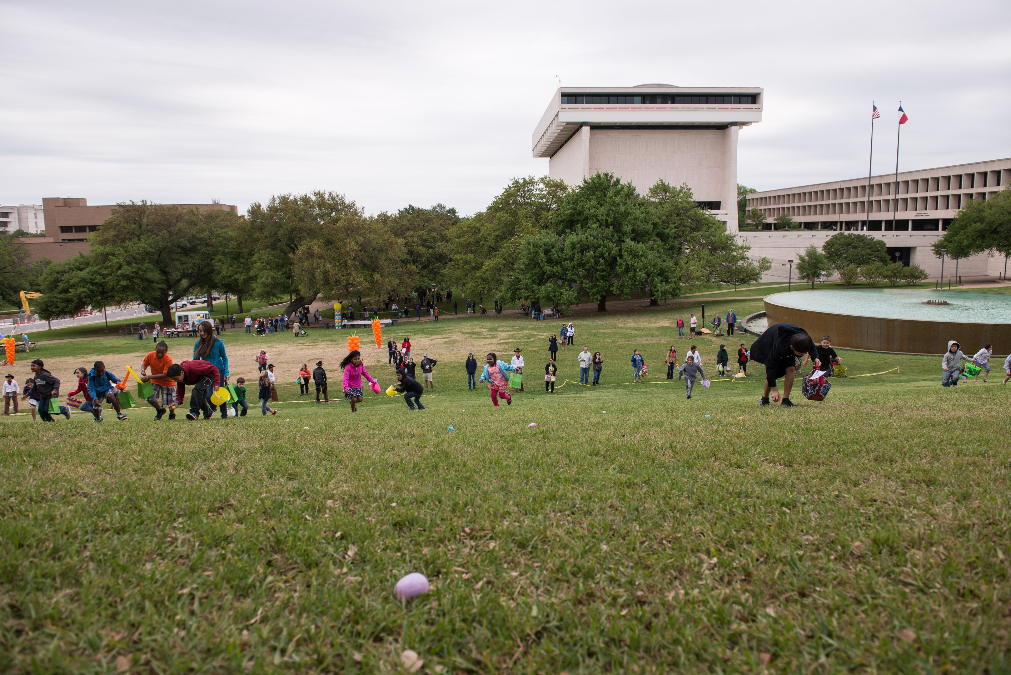 Gruppe von Menschen beim Ostereier-Spiel auf einer grünen Wiese mit Bäumen, Gebäuden, Fahnen, einem Brunnen und einem bewölkten Himmel im Hintergrund.