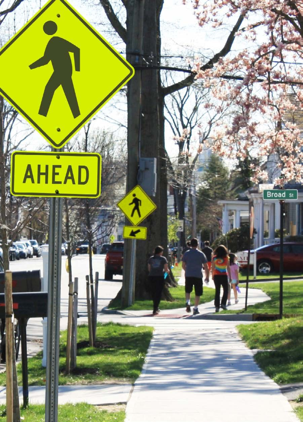 Eine Gruppe von Menschen, die auf einem Gehweg neben einem gelben Fußgängerübergangsschild geht, mit Fahrzeugen auf der Straße, Gebäuden im Hintergrund und einem klaren blauen Himmel.