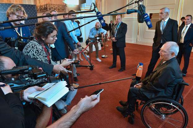 Ein Mann im Anzug und Krawatte sitzt im Rollstuhl, umgeben von Reportern mit Kameras und Mikrofonen sowie einem Banner und einer Wand im Hintergrund.