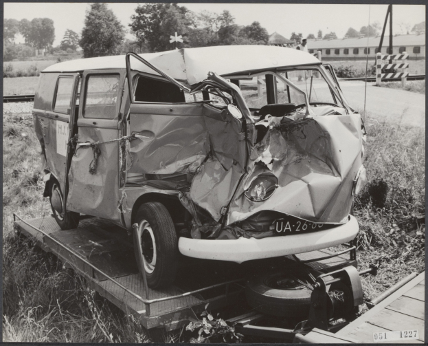 Ein schwerbeschädigtes schwarzes Auto mit eingedellter Front und zersplitterten Fenstern auf einem Tieflader, umgeben von Gras und Pflanzen, mit Bäumen und einem Gebäude im Hintergrund.