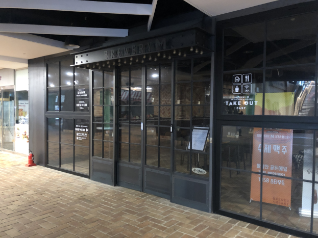 Der Eingang zu einem Gebäude mit Glaswänden und -türen, ein Schild mit der Aufschrift "Brauerei M Stable", ein Feuerhydrant auf dem Boden und Deckenleuchten.