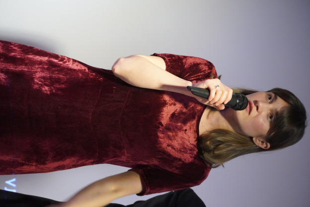 Eine Frau in einem roten Kleid steht selbstbewusst auf einer Bühne, hält ein Mikrofon in der Hand und hat eine stylische Bob-Frisur sowie natürliches Make-up.