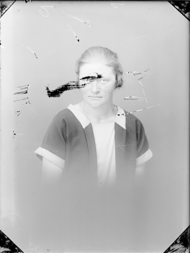 Ein Schwarz-Weiß-Foto einer Frau mit einem zerbrochenen Glasfenster im Hintergrund, ihr Gesicht ist deutlich zu sehen, mit direktem Augenkontakt und leicht geöffneten Lippen, das Haar zurückgekämmt und die Hände vor der Brust verschränkt.