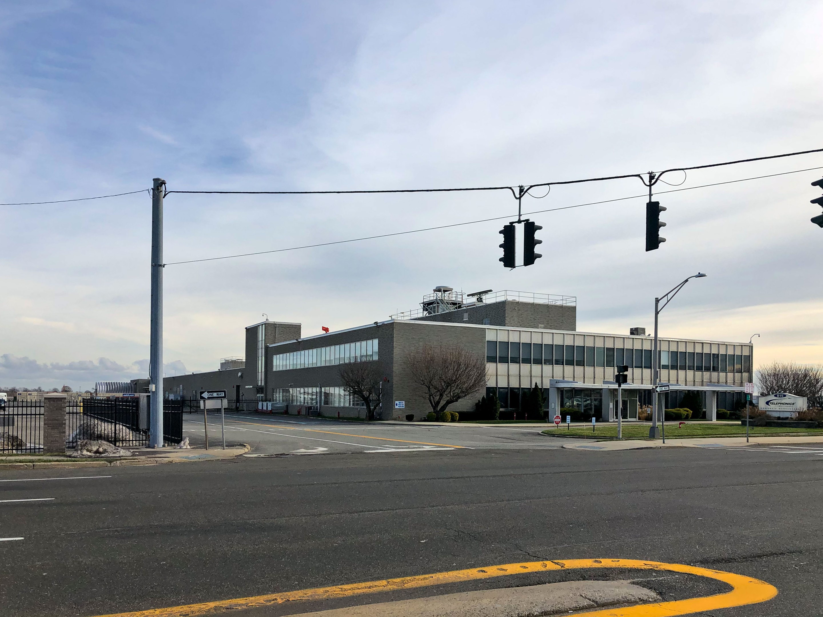 Großes Gebäude mit zahlreichen Verkehrsampeln, Straßenlaternen und elektrischer Infrastruktur, umgeben von Kraftfahrzeugen, Bäumen und einem bewölkten Himmel, als die neue Hauptquartier der Nationalen Sicherheitsbehörde identifiziert.