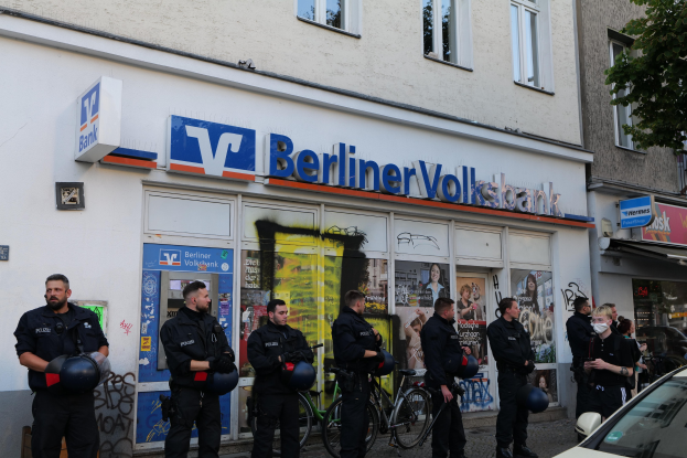 Polizeibeamte vor einer Berliner Volksbank, einige mit Helmen, daneben ein Auto und ein Baum, im Hintergrund Gebäude mit Fenstern und Texttafeln.