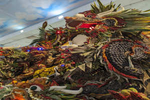 Ein großer Haufen Obst und Gemüse in einem Raum mit beleuchteter Decke und einer Wand im Hintergrund, mit dem Text 'Papua-Neuguinea-Karneval' unten.