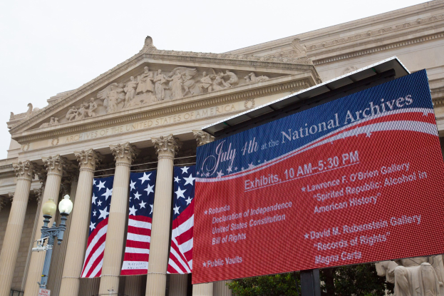 Übersicht des Nationalarchiv-Gebäudes in Washington, DC, mit einer Fahne, Flaggen, Leuchten, Bäumen und Skulpturen unter einem klaren blauen Himmel.
