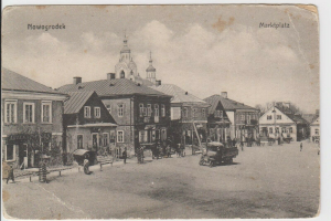 Schwarze und Weiße Fotografie einer Straße in Nowogrodziek, Deutschland, mit Gebäuden, Menschen, Wagen, Pfölen, Bäumen und Himmel, mit der Aufschrift "Marktplatz in Nowogrodeek, Deutschland" oben und unten.
