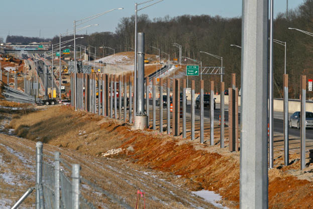 Baustelle mit Fahrzeugen, Pfählen, Beleuchtung, Beschilderung, einem Zaun, schneebedeckter Wiese, Bäumen und einem Himmel im Hintergrund.