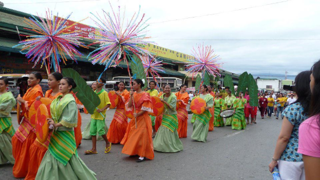 Eine Gruppe von Menschen in farbenfrohen Kostümen geht eine Straße entlang während eines Karnevalsumzugs, mit einer Frau auf der rechten Seite, die eine Flasche hält, und Fahrzeugen, Gebäuden, Bäumen, Pfählen, Drähten und dem Himmel im Hintergrund.
