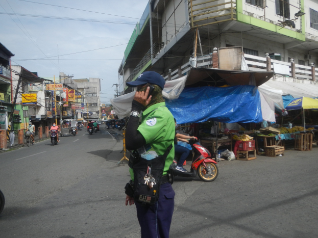 Polizist in grüner und blauer Uniform und Mötte, der auf einer Straße steht und mit dem Handy spricht, mit Fahrzeugen und Marktbuden im Hintergrund und einem Gebäude am bewölkten Himmel.