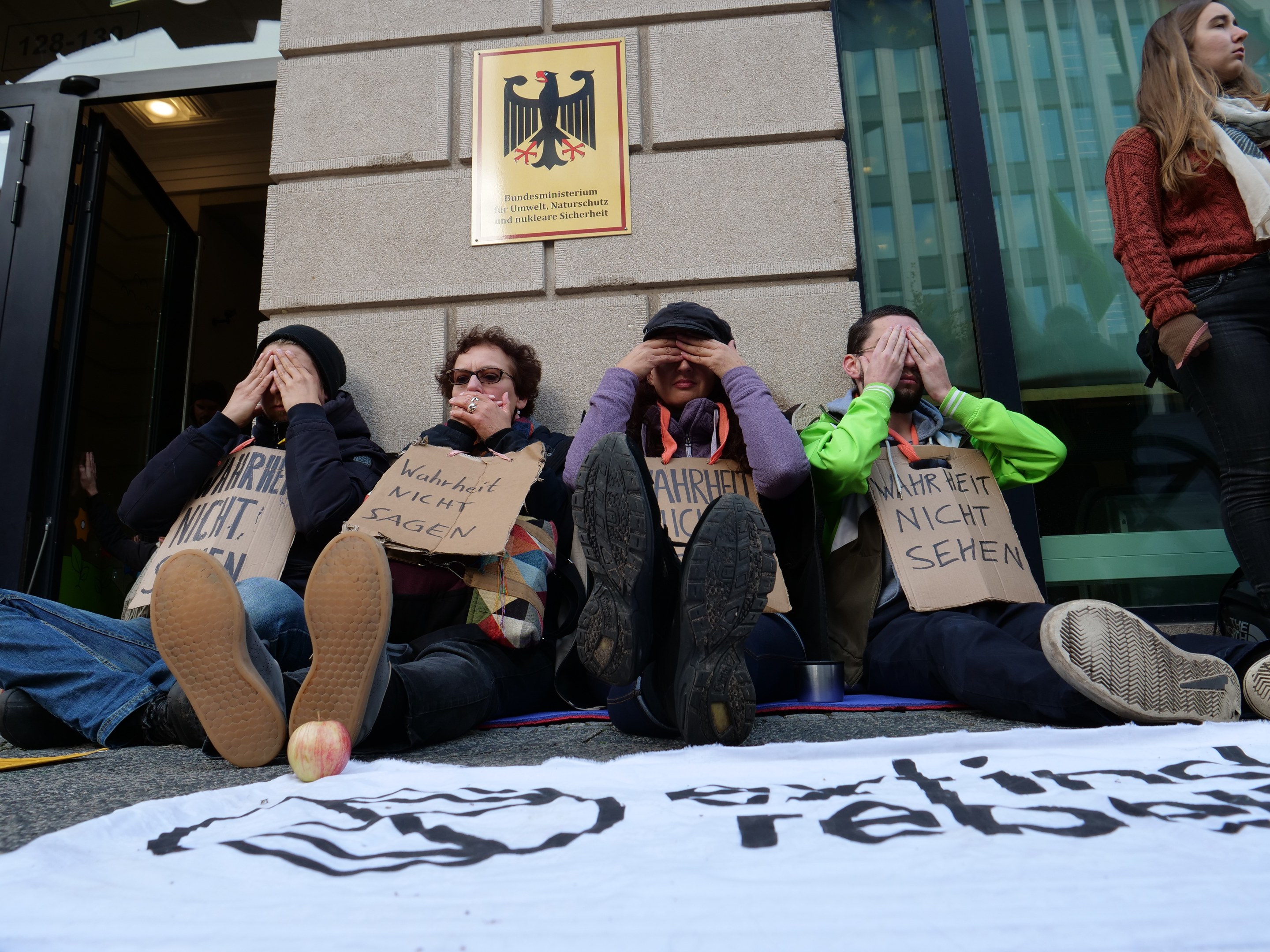 Eine Gruppe von Menschen sitzt vor einem Gebäude auf dem Boden und hält Protestschilder mit Text, mit einem Banner auf der Straße und einer Frau, die rechts steht, vor einer Wand mit einer Tafel und einem Gebäude mit Glasfenstern und einer Tür.