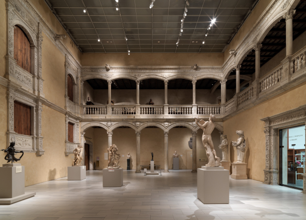 Ein großer Museumsgalerie mit zahlreichen Statuen und Skulpturen auf Sockeln, architektonischen Elementen wie Säulen und Geländern, Deckenbeleuchtung und einigen Besuchern.