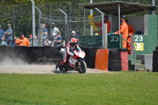 Ein Motorradfahrer mit Helm und Handschuhen fährt über ein grasbewachsenes Feld, mit Zuschauern in Mützen und Schutzbrillen, einem eingezäunten Bereich, Treppen mit Geländern und einem nummerierten Schuppen im Hintergrund.
