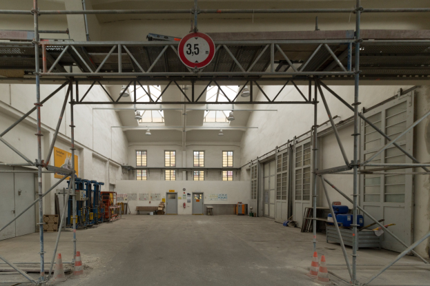 Große Lagerhalle mit ausgedehnten Gerüten, Verkehrskegeln, Metallstangen, einem Schild, Maschinen, einer Tür, Fenstern, Wänden, Deckenlampen und Dach.