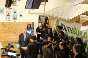 Eine Gruppe von Menschen steht um ein Rednerpult vor einer Menge, mit einem Tisch mit Mikros und Papieren links, Plakaten an der Wand im Hintergrund, Gegenständen auf dem Dach und einem Gegenstand rechts, Händeschütteln, was darauf hinweist, dass sie Teil einer Jugendgruppe sind.