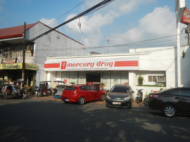Eine belebte Straßenszene mit einer Mercury Drug Store im Vordergrund, mit Fahrzeugen auf der Straße, Gebäuden mit Namensschildern, Strommasten mit Drähten und einem bewölkten Himmel im Hintergrund.