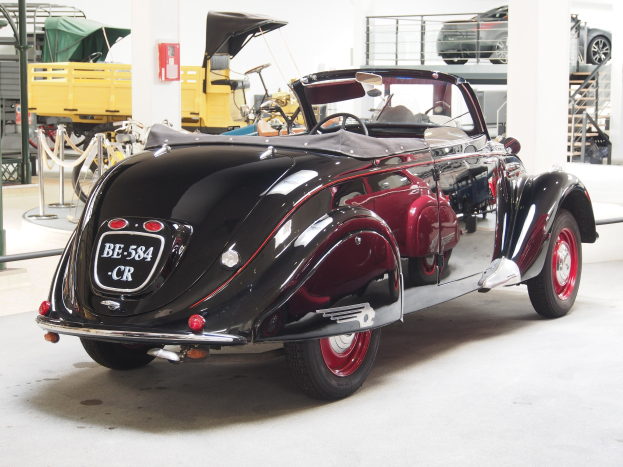 Ein schwarzes und rotes Auto in einer Garage mit anderen Fahrzeugen, Pfosten, Ketten und Gegenständen geparkt, mit einem Feuerlöscher an der Wand im Hintergrund.