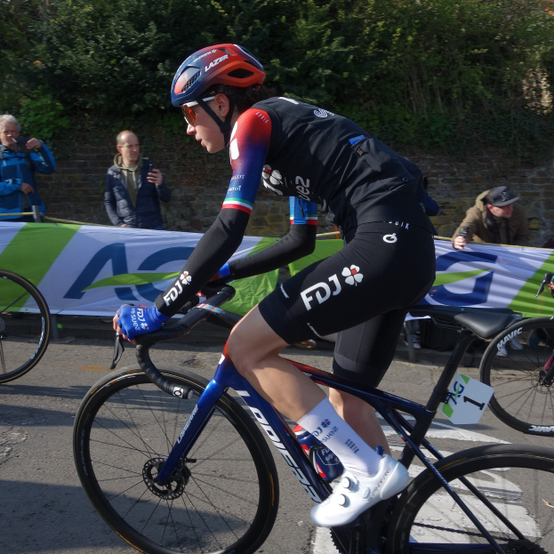 Professionelle Radfahrerin Katie Archibald trägt einen Helm, eine Brille, Handschuhe und Schuhe und fährt ein Fahrrad auf einer Straße neben einer Menge, mit einem Banner, einer Wand, Bäumen und einem Gebäude im Hintergrund.