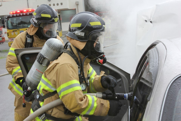 Feuerwehrleute in Schutzausrüstung löschen ein brennendes Auto mit einem Schlauch, während Rauch aus dem Fahrzeug aufsteigt.