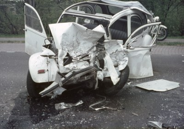 Ein schwer besch├Ądigtwagen mit Frontalaufprall am Stra├čenrand, umgeben von B├Ąumen und einem Geb├Ąude im Hintergrund, mit einem weiteren Fahrzeug in der Ferne.