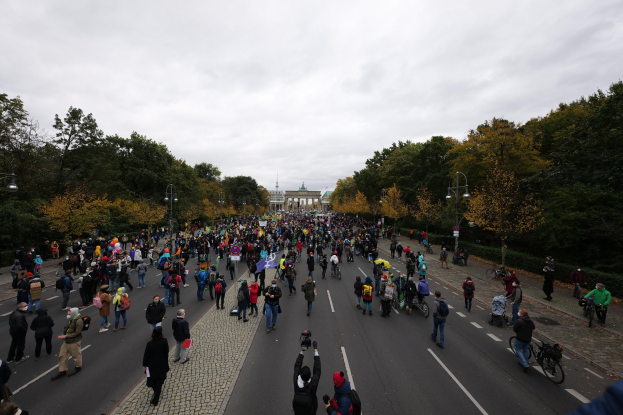 Eine große Gruppe von Menschen marschiert auf einer von Bäumen gesäumten Straße mit Laternenmasten, mit Kameras in der Hand, mit einem Gebäude und einem klaren Himmel im Hintergrund, was auf eine Demonstration in Berlin hinweist.