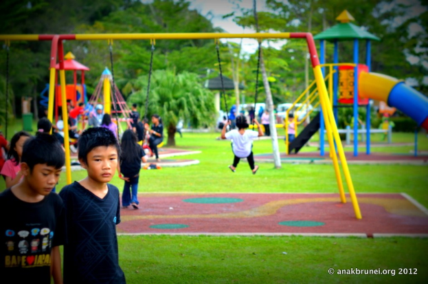 Kinder auf Spielgeräten in einem Park mit Bäumen im Hintergrund.