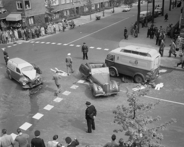 Schwarz-weißes Bild einer Autounfall-Szene an einer Straßenkreuzung mit mehreren Fahrzeugen, darunter ein Lieferwagen, und einer Gruppe von Zuschauern; Gebäude, Bäume, Pfosten und Texttafeln im Hintergrund.