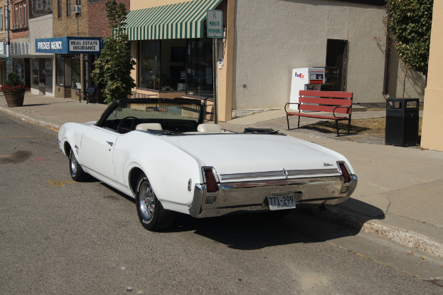 Ein weißes Auto mit der Nummer 771-299 steht auf einer Straße, daneben eine Bank und im Hintergrund ein Gebäude mit einem "Immobilienversicherung"-Geschäft.