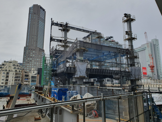 Großer Bauplatz in einer Stadt mit einem teilweise errichteten Kraftwerk, umgeben von einem Metallzaun, Fahrzeugen, Masten, einer Plane, näheren Gebäuden und einem bewölktem Himmel.