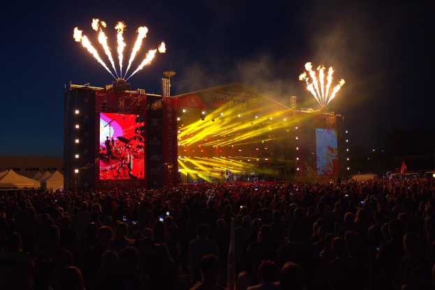 Eine große Menschenmenge auf einem Musikfestival mit einer Bühne, auf der Künstler unter hellen Scheinwerfern spielen, umgeben von Bannern, Zelten links und Feuerwerk am dunklen Himmel dahinter.