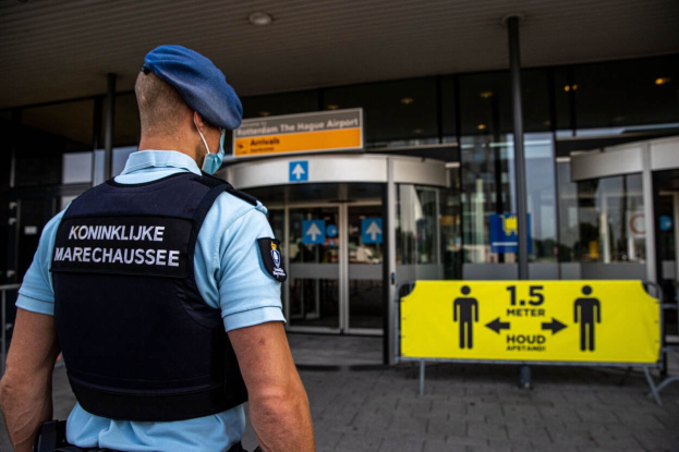Ein Polizist in blauer Uniform und Maske steht vor einem Glasgebäude, mit gelben und anderen Schildern im Hintergrund.