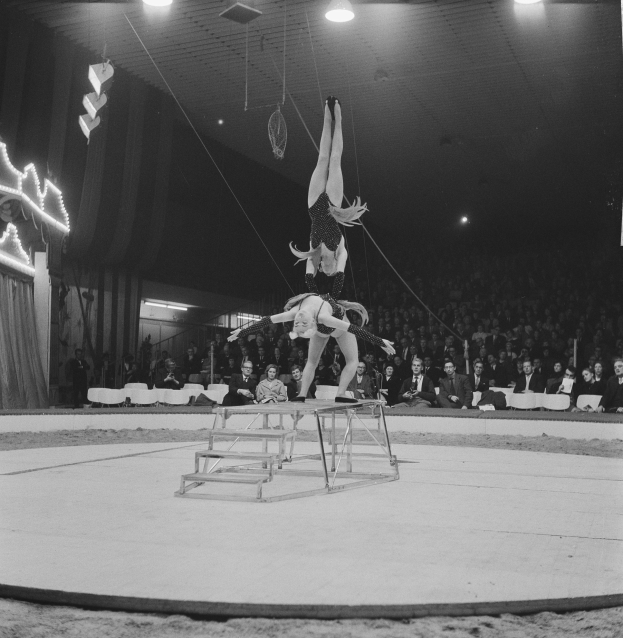 Ein Schwarz-Wei-Foto von zwei Akrobaten, die auf einer von Deckenleuchten beleuchteten Bühne Kunststücke zeigen, vor einem sitzenden Publikum.