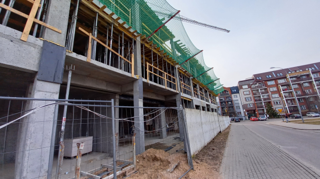 Großes Bauprojekt an einer Stadtkreuzung mit Straßenlaternen, Lampen, Fahrzeugen und umliegenden Gebäuden unter einem klaren blauen Himmel.