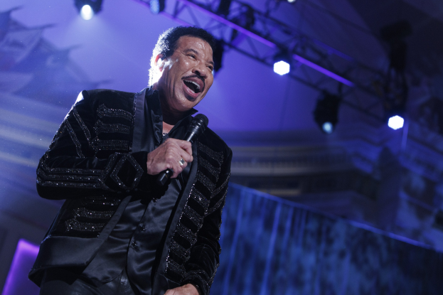 Lionel Richie performs bei den Grammys, trägt ein schwarzes Kleid und hält ein Mikrofon in der Hand, während helle Bühnenlichter eine dramatische Atmosphäre schaffen.