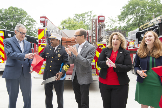 Gruppe von Menschen bei einer Eröffnungszeremonie für die Alexandria Fire Department, die vor einem Feuerwehrauto klatschen und lächeln, mit Bäumen und einem blauen Himmel im Hintergrund.