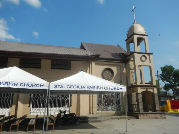 St. Cecilia Parish Church in Cebu City, Philippines, with a clock on its wall, surrounded by trees, utility poles, wires, a tent, and a parked motorbike under a cloudy sky.