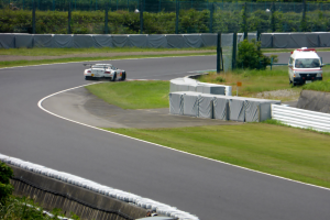 Ein Rennwagen fährt auf einer Rennstrecke mit einem Polizeiwagen im Hintergrund, umgeben von Vegetation und Absperrungen, mit Text am unteren Bildrand.
