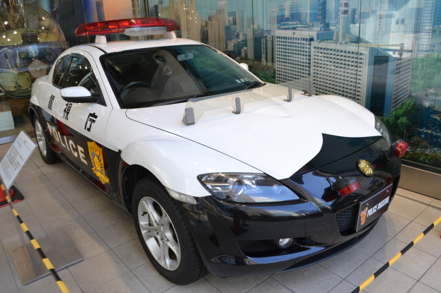 Ein Polizeiwagen steht vor einer Glaswand, mit einem sichtbaren Rad, auf einem Boden neben einer Tafel mit Text. Im Hintergrund sind durch die Glaswand Gebäude, Bäume und der Himmel zu sehen.