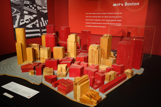 Ein hölzernes Blockmodell einer Stadt, das auf einem Tisch in einem Museum ausgestellt ist, begleitet von Papieren, einer Glasbox und einem Rahmenfoto an der Wand.