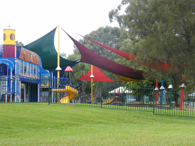 Ein Park mit einem eingezäunten Spielplatz mit einer Rutsche, Stangen und einem Zelt, auf dem Kinder spielen, umgeben von grünem Gras, Bäumen und einem klaren blauen Himmel.