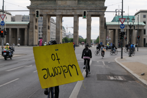 Eine Gruppe von Menschen fährt auf Fahrrädern eine Straße entlang und passiert das Brandenburger Tor in Berlin, Deutschland, mit Helmen, wobei eine Person ein gelbes Schild hält, unter einem klaren blauen Himmel mit Gebäuden und Bäumen im Hintergrund.
