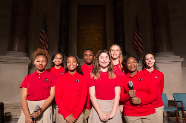 Gruppe junger Frauen in roten Shirts, die strahlend Mikrofone halten und vor Stühlen, Säulen, Flaggen und einer Wand in einem Saal stehen.