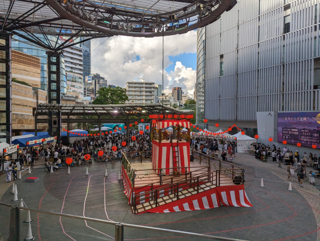 Große Menschenmenge um eine Bühne in einer städtischen Umgebung mit Gebäuden, Bäumen und einem bewölkten Himmel, mit Performern und Equipment für die Eröffnung des Tokyo Circus Festivals.