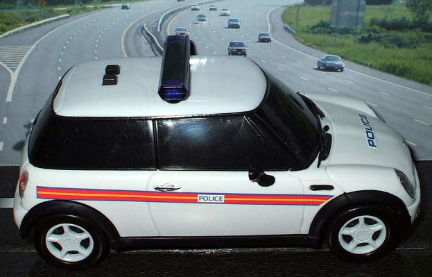 Weißer Polizeiwagen mit "Polizei"-Aufschrift auf einer Landstraße außerhalb der Stadt, umgeben von fließendem Verkehr, Gras und Bäumen auf beiden Seiten.