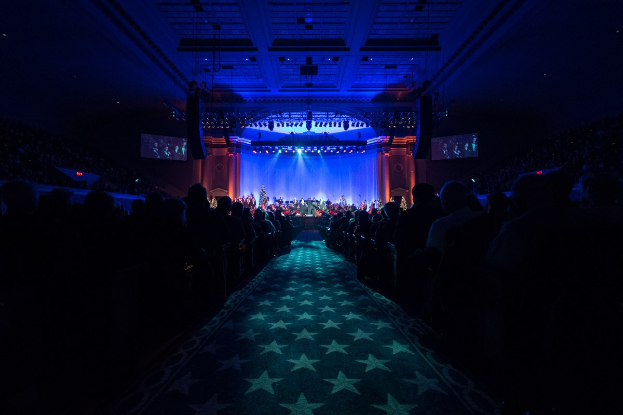 Ein großer Zuschauerraum mit Sitzplätzen, eine Bühne mit Musikern und Deckenleuchten und -fernseher, was auf einen Konzertsaal während der Nationalkonvention 2019 hindeutet.